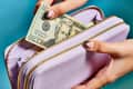 Hands placing a $20 bill into a lavender wallet with gold zippers against a blue background.