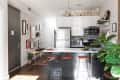 Modern kitchen with white cabinets, stainless steel appliances, bar stools, and a plant. Wall art decorates the adjacent wall.