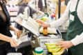 Cashier in green apron scanning bananas at grocery checkout counter.