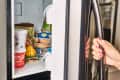 Hand with red nails opening a freezer door, revealing ice cream, frozen vegetables, and packaged food inside.