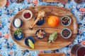Wooden platter with an orange, bone, egg, artichoke, olives, nuts, and small bowls on a floral tablecloth.