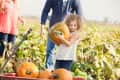 Child holding a pumpkin in a field, with adults nearby and a wagon filled with pumpkins.