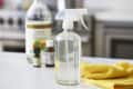 Spray bottle with cleaning solution, vinegar, essential oils, and yellow cloth on a kitchen counter.
