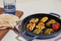 Grilled chicken and vegetables in a blue pan with pita bread and a glass of water on a wooden board.