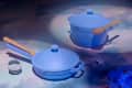 Blue cookware set with wooden handles, including a pot and pan, on a cloud-patterned surface with glass spheres.