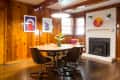 Dining room with wooden walls, round table, four chairs, white brick fireplace, and colorful wall art.