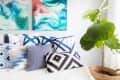 Bed with assorted blue and white patterned pillows, abstract paintings above, and a large potted fiddle leaf fig plant.