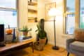 Cozy living room with a gray armchair, floor lamp, cactus, and wall art. Coffee table with books, plants, and a pumpkin.