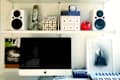 Home office with iMac, keyboard, speakers, microphone, and shelves with books, boxes, and decor items.