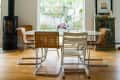 Dining room with wicker chairs, round table, pendant light, and large window overlooking a garden.
