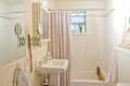 Small bathroom with white tiles, pedestal sink, extendable mirror, and bathtub with beige curtain and wooden bath tray.