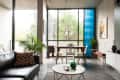 Modern living room with large windows, black leather sofa, dining table, plants, and abstract wall art.