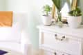 White dresser with potted plants, a mirror, and a framed picture on a light green wall.