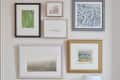 Gallery wall with various framed artworks above a bar cart with bottles, a gold bowl, and a white lamp.