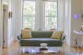 Living room with a green sofa, yellow pillows, floral chandelier, and large windows overlooking trees.