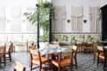 Restaurant interior with macramé wall hangings, patterned floor tiles, wooden chairs, and floral cushioned seating.