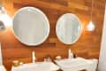 Two white sinks with round mirrors on a wooden wall, pendant lights, and toiletries in a modern bathroom.