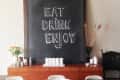 Chalkboard with "Eat Drink Enjoy" above a wooden sideboard, vase of flowers, and a bowl on a dark dining table.