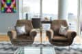 Two brown armchairs with floral pillows in a modern living room, large windows, colorful abstract art, and a glass coffee table.