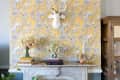 Yellow floral wallpaper with a white deer head, marble mantel, books, vases of flowers, and brass candlesticks.