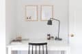 Minimalist workspace with a white desk, black chair, desk lamp, and framed art on the wall.