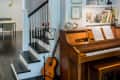 Wooden piano with sheet music, guitar, and music stand beside a staircase in a cozy living room.