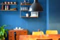 Dining area with wooden table, mixed chairs, blue wall, shelves with dishes, and a large black pendant light.