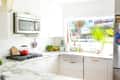 Bright kitchen with marble countertops, red kettle on stove, potted plants, and large window with palm leaf view.