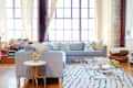 Loft living room with a gray sectional sofa, colorful pillows, glass coffee table, and stacks of books by large windows.