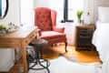 Bedroom corner with a coral armchair, wooden desk, black stool, and cowhide rug near a window with white curtains.