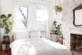 Bright bedroom with white bedding, brick fireplace, hanging plants, and large windows with sheer curtains.