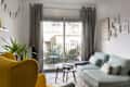 Cozy living room with a yellow armchair, light blue sofa, indoor plant, and wall art, facing a balcony with outdoor seating.