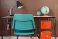 Teal chair at a desk with a black lamp, globes, orange boxes, and a colorful abacus on a brown wall.