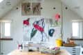 Sewing room with a wooden table, sewing machine, fabric stacks, and colorful artwork on the wall.