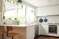 Modern kitchen with white cabinets, wooden stools, hanging plants, and a large window with striped awning.