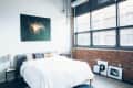 Loft bedroom with a large window, brick wall, modern art, and a bed with a white comforter.