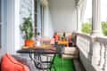 Balcony with metal table, orange cushions, potted plants, and decorative pillows on a green artificial grass rug.