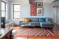 Living room with gray sectional sofa, colorful pillows, vibrant patterned rug, and framed artwork on light blue walls.