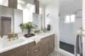 Modern bathroom with dual sinks, gold fixtures, wood cabinets, large mirrors, and a glass shower.