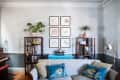 Living room with a gray sofa, blue peacock pillows, wooden shelves, plants, and framed art on a light gray wall.
