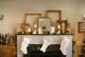 Fireplace mantel decorated with gold frames, candles, and white stockings, surrounded by festive greenery and bottle collection.