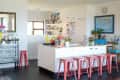 Modern kitchen with white island, pink stools, hanging lights, and colorful decor, including a world map and potted plants.