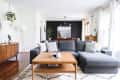 Modern living room with gray sectional sofa, wooden coffee table, and plants, leading to a dining area with dark accent wall.