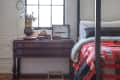 Loft bedroom with a four-poster bed, red and black quilt, wooden desk, and large window with a wall-mounted lamp.