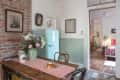 Rustic kitchen with a wooden table, pink runner, flowers, pastries, and a mint green retro fridge. Bedroom visible through doorway.