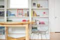 Wooden desk with wire chairs in front of white bookshelves filled with colorful books and decorative items.