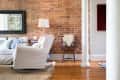 Living room with brick wall, white armchair, blue patterned pillows, floor lamp, and a wooden chair with a fur throw.