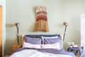Bed with gray and white pillows, wall-mounted lamps, and woven wall art on a light green wall.