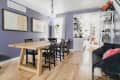 Dining room with a wooden table, black chairs, purple walls, and framed art, leading to a bright kitchen with plants.