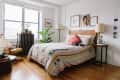 Cozy bedroom with a wooden bed, geometric quilt, pink pillow, and a cat. Plants and framed art decorate the space.